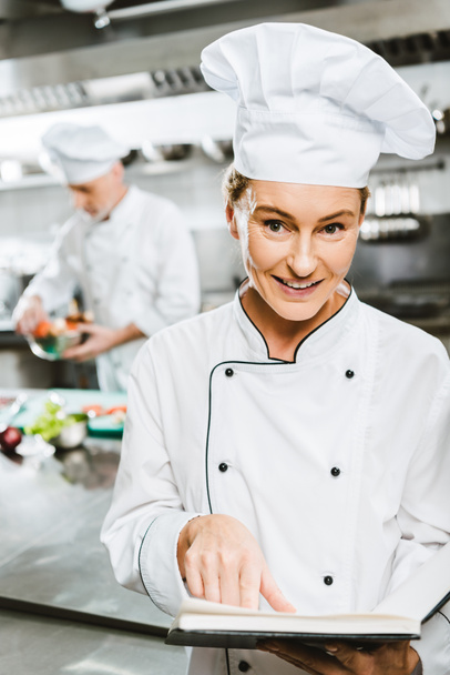 belle femme chef en uniforme tenant livre de recettes et regardant caméra dans la cuisine du restaurant
 - Photo, image