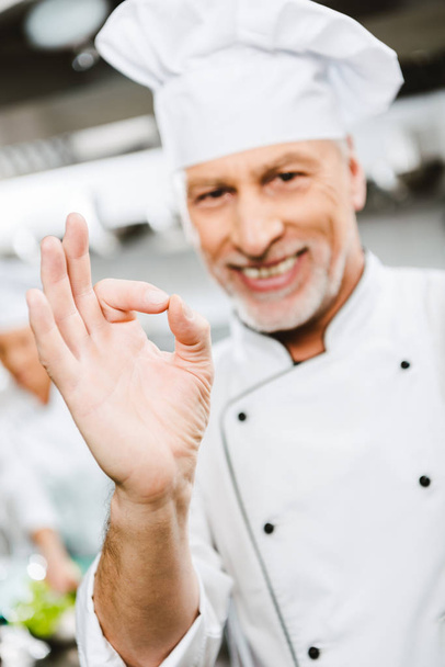 handsome smiling male chef in uniform looking at camera and showing ok sign in restaurant kitchen - Fotó, kép