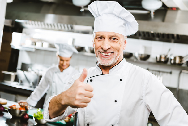 レストランのキッチンでハンサムな制服を着た男性シェフの笑みを浮かべてカメラを見て、親指を現してサインします。 - 写真・画像