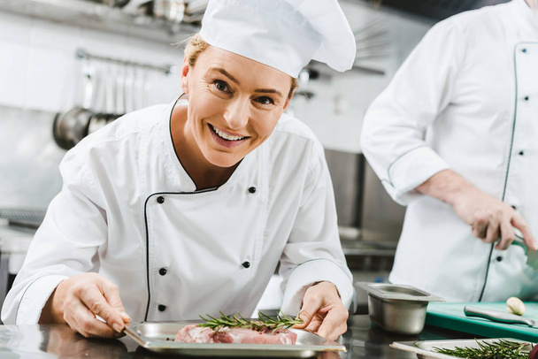 beautiful female chef in uniform holding plate with meat dish and looking at camera in restaurant kitchen - Photo, Image