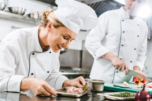 lächelnde Köchin mit Fleischgericht, während Kollegin in Uniform im Hintergrund in der Restaurantküche kocht - Foto, Bild