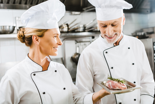cocinera sonriente en uniforme presentando plato de carne a colega en la cocina del restaurante
 - Foto, imagen