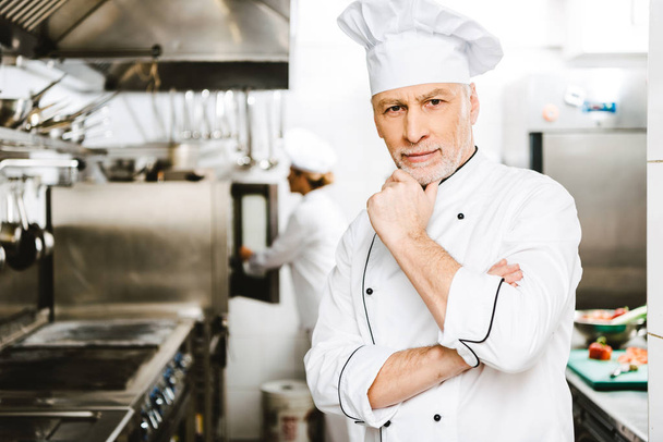 handsome male chef propping chin with hand and looking at camera in restaurant kitchen - Foto, afbeelding