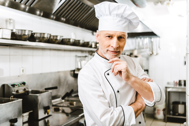 handsome male chef in uniform and cap touching chin andf looking at camera in restaurant kitchen - Foto, imagen