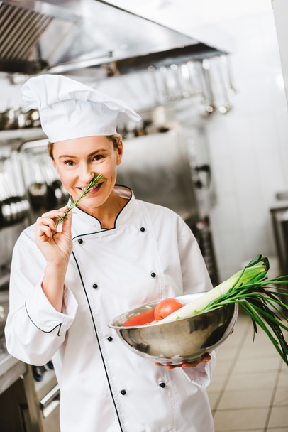 beautiful smiling female chef in uniform holding rosemary and bowl with vegetables in restaurant kitchen - Photo, Image