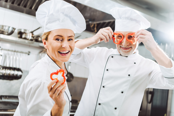 Szczęśliwa kobieta kucharz patrząc na kamery, podczas gdy mężczyzna kucharz trzyma pieprz plastry przed twarzą w kuchni restauracji - Zdjęcie, obraz