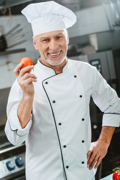 usmívající se muž kuchař v uniformě a klobouk drží rajče a při pohledu na fotoaparát v kuchyni restaurace - Fotografie, Obrázek