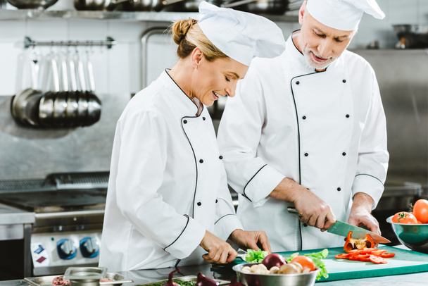 chefs femeninos y masculinos sonrientes en uniforme cortando ingredientes mientras cocinan en la cocina del restaurante
 - Foto, imagen