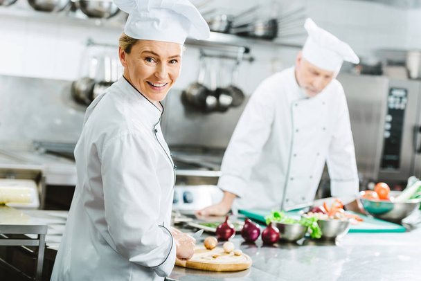 piękna kobieta kucharz patrząc na kamery i uśmiechając się podczas gotowania z kolega w kuchni restauracji - Zdjęcie, obraz
