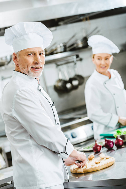 handsome male chef looking at camera while cooking with female colleague in restaurant kitchen - 写真・画像