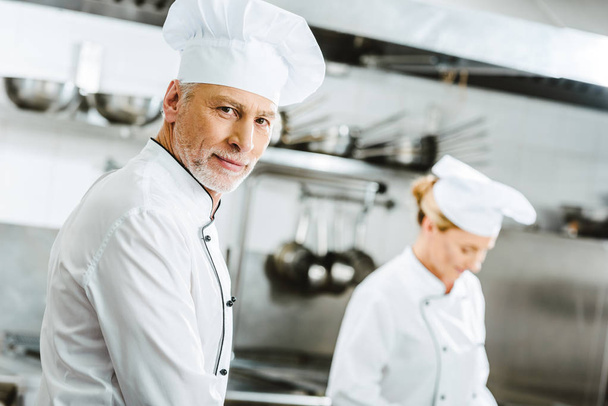 valikoiva painopiste komea mies kokki kaksirivinen takki ja hattu katselee kameraa ravintolan keittiössä
 - Valokuva, kuva