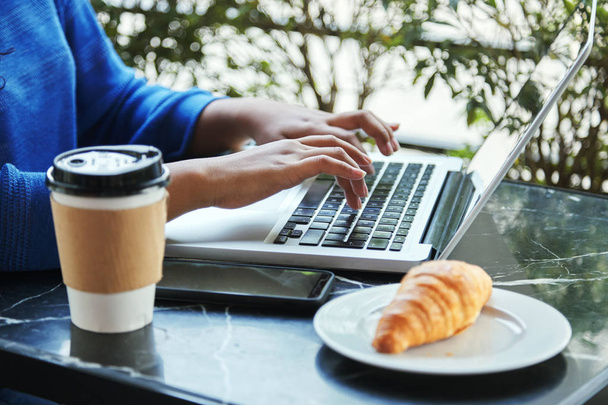 カフェのテーブルとクロワッサン近くのノート パソコンのキーボードで入力してホットド リンクのカップで座っている女性の作物ショット - 写真・画像