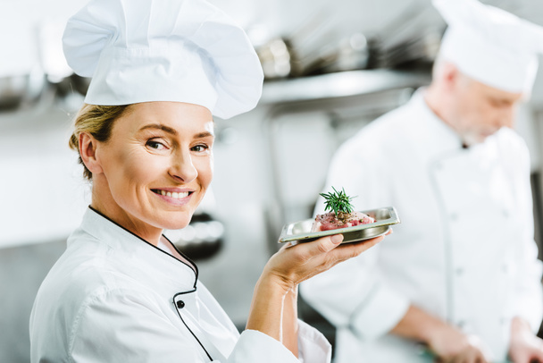 beautiful smiling female chef in uniform holding plate with meat dish in restaurant kitchen and looking at camera - Фото, изображение