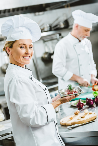 female chef in uniform holding meat dish on plate with male colleague cooking on background in restaurant kitchen - Foto, imagen