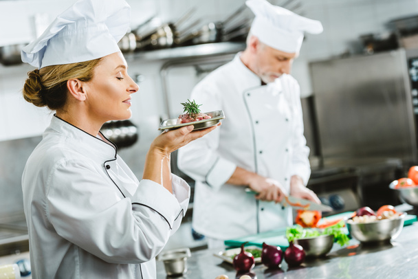 chef feminino em uniforme segurando prato de carne na placa com colega masculino cozinhar no fundo na cozinha do restaurante
 - Foto, Imagem