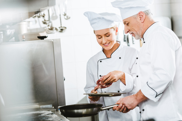 ženské a mužské kuchaři v uniformě vaření masa na pánvi v kuchyni restaurace s kopií prostor - Fotografie, Obrázek
