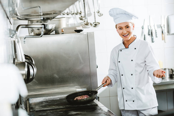 beautiful smiling female chef in uniform looking at camera while cooking meat in restaurant kitchen - Photo, Image