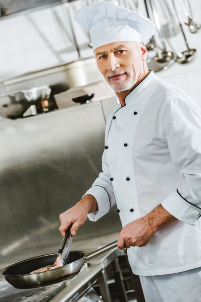 male chef looking at camera while roasting meat steak in restaurant kitchen - Photo, Image