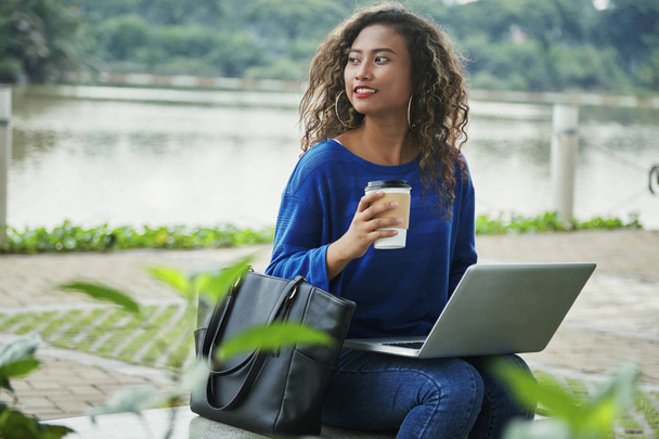 Pretty Indonesian woman in casual outfit smiling and looking away while sitting on city street near water and holding cup of hot drink and modern laptop - Photo, image