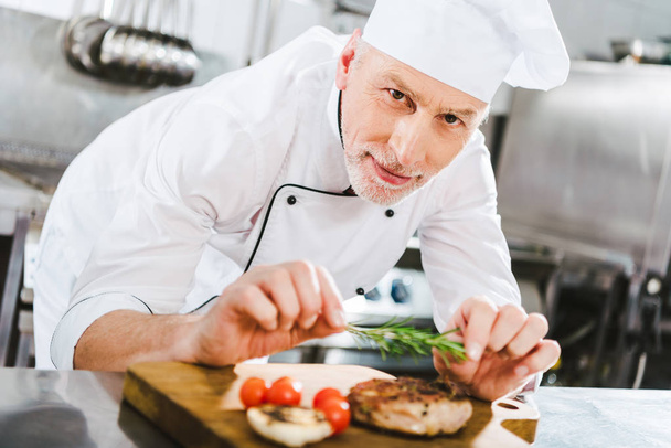 chef masculin en uniforme décorant steak de viande avec romarin et regardant la caméra dans la cuisine du restaurant
 - Photo, image