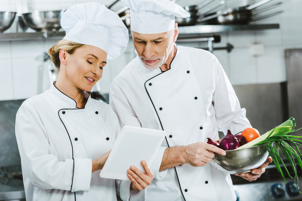 жіночі та чоловічі шеф-кухарі у формі проб використовують цифровий планшет під час приготування їжі на кухні ресторану
 - Фото, зображення