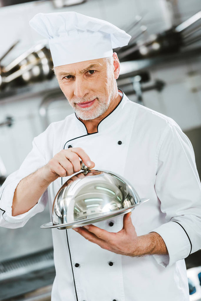 beau chef masculin en uniforme regardant la caméra et tenant plateau de service avec dôme dans la cuisine du restaurant
 - Photo, image