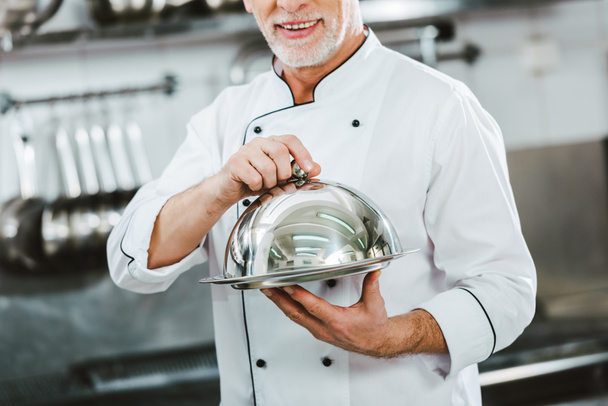 περικοπεί άποψη των αρσενικών σεφ σε ομοιόμορφη εκμετάλλευση δίσκο σερβιρίσματος με θόλο σε κουζίνα εστιατορίου - Φωτογραφία, εικόνα