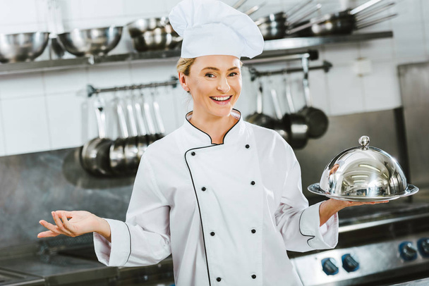 レストランのキッチンでドームのサービング トレイを保持している制服を着た美しい笑みを浮かべて女性シェフ - 写真・画像