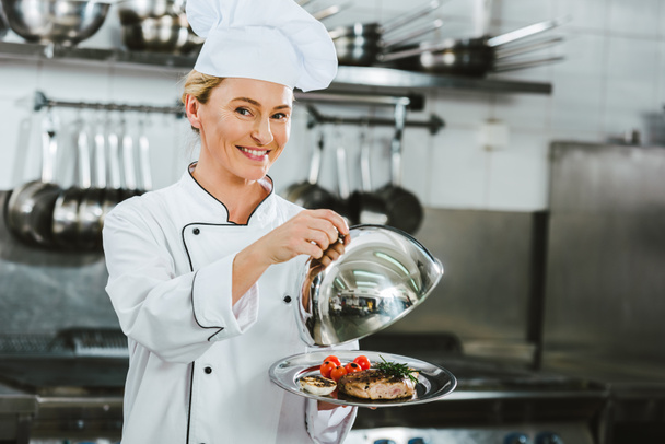hermosa mujer sonriente chef en uniforme de la celebración de la cúpula de servir bandeja con plato de carne en la cocina del restaurante
 - Foto, Imagen