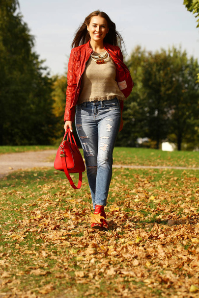 Young beautiful woman in fashion red jacket and blue jeans walking in autumn park - Fotoğraf, Görsel