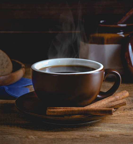 Steaming cup of coffee with cinnamon sticks on a wooden surface in a dark rustic setting - Foto, imagen