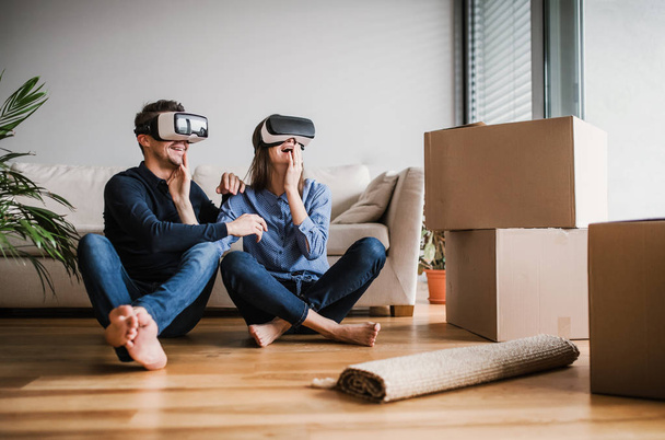 A young couple with VR goggles sitting on a floor, moving in a new home. - Foto, Imagen