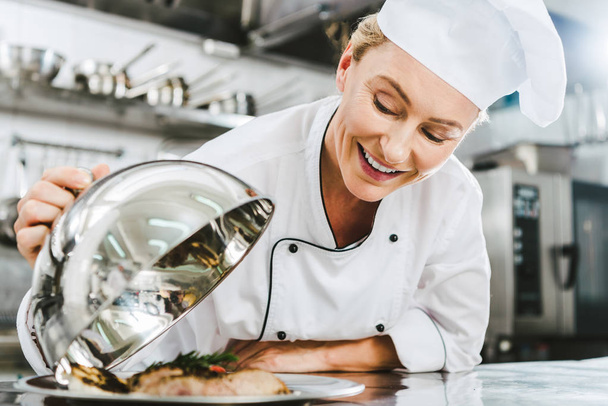 beautiful female chef in uniform holding dome from serving tray with meat dish in restaurant kitchen - Foto, Bild