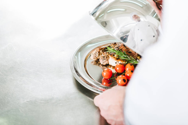 vista parcial del chef masculino sosteniendo la cúpula de servir bandeja con plato de carne en la cocina del restaurante
 - Foto, imagen