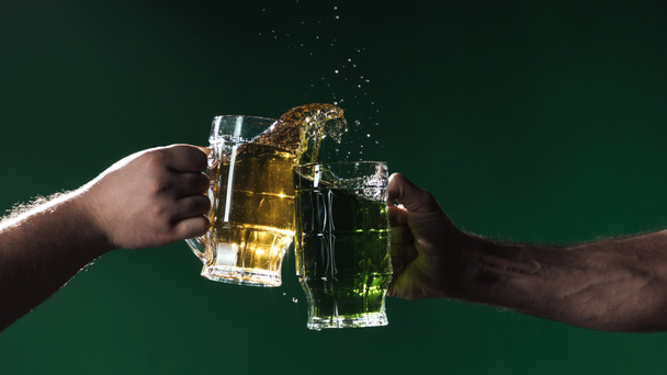 vista parziale degli uomini che si tintinnano bicchieri di birra con spruzzi isolati sul verde scuro, concetto di giorno di San Patrizio
 - Foto, immagini