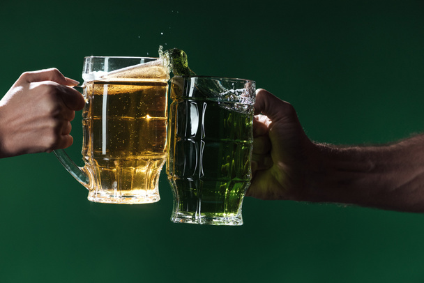 partial view of men clinking glasses of beer with splashes isolated on dark green, st patrick day concept - Foto, Imagen