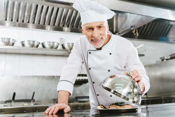 όμορφος χαμογελώντας αρσενικό σεφ σε ομοιόμορφη εκμετάλλευση θόλου από δίσκος σερβιρίσματος με πιάτο κρέατος σε κουζίνα εστιατορίου - Φωτογραφία, εικόνα