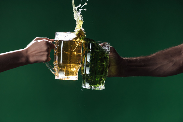 abgeschnittene Ansicht von Männern, die Biergläser mit Spritzern auf dunkelgrünem, st patrick day Konzept klirren - Foto, Bild