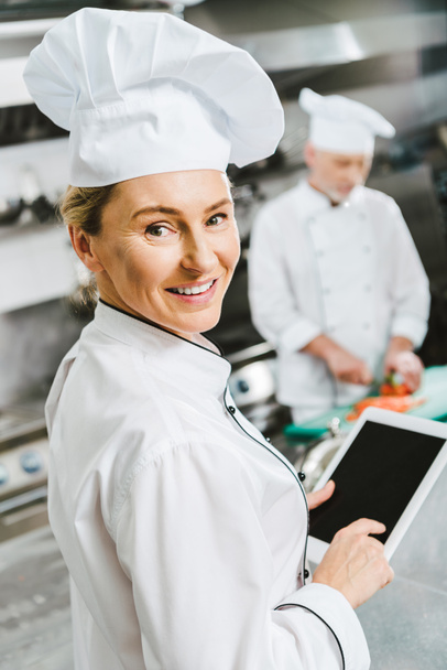 enfoque selectivo de la mujer chef sonriente mirando a la cámara y el uso de tabletas digitales con pantalla en blanco en la cocina del restaurante
 - Foto, Imagen