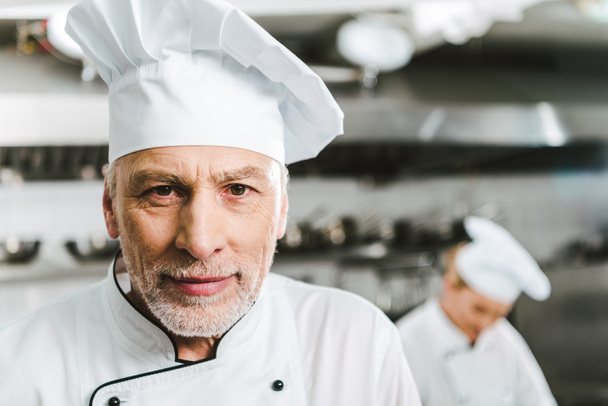 knappe mannelijke chef-kok in uniform en cap camera in restaurant keuken kijken met kopie ruimte - Foto, afbeelding