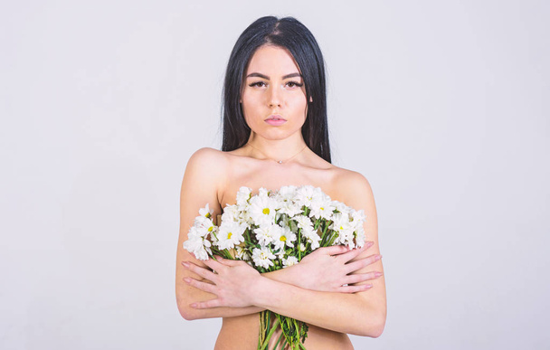 Girl on calm face stands naked and holds chamomile flowers in front of chest. Lady covers breasts with flowers, isolated on grey. Skin care concept. Woman with smooth healthy skin looks attractive - Photo, Image