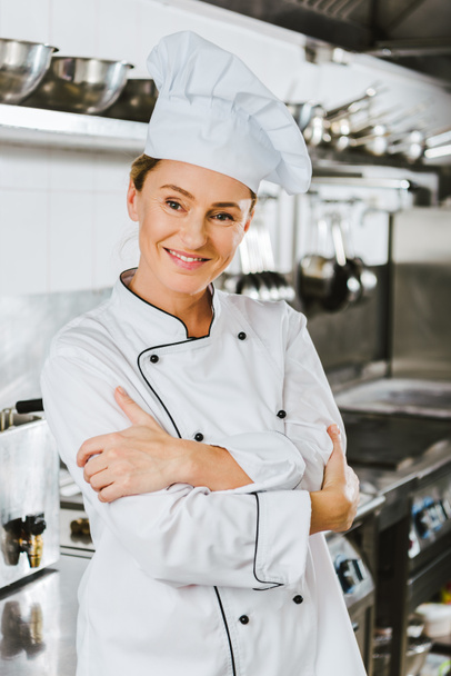 attractive female chef in uniform with arms crossed looking at camera in restaurant kitchen - Fotó, kép