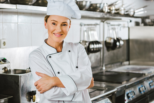 atractiva mujer chef en uniforme con los brazos cruzados mirando a la cámara en la cocina del restaurante
 - Foto, Imagen