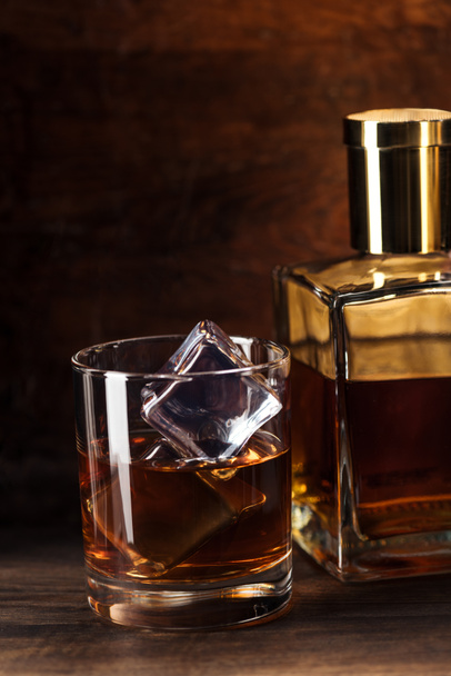 close-up view of glass of brandy with ice cubes and bottle on wooden table   - Foto, Imagen