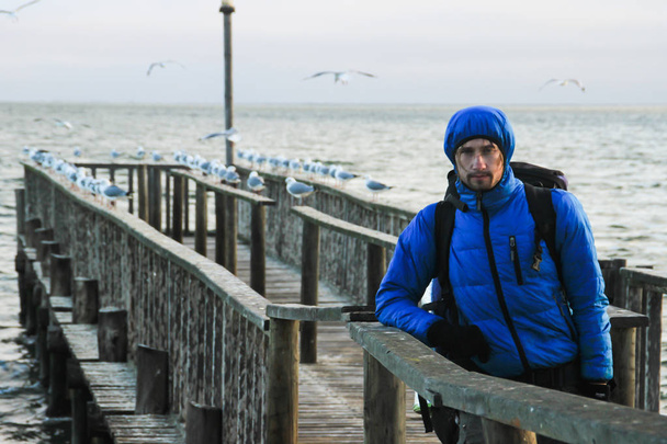 White Caucasian male traveler in a sports blue jacket, gloves, hat, glasses and a backpack standing on the pier of the Atlantic Ocean, surrounded by sea gulls - Fotografie, Obrázek