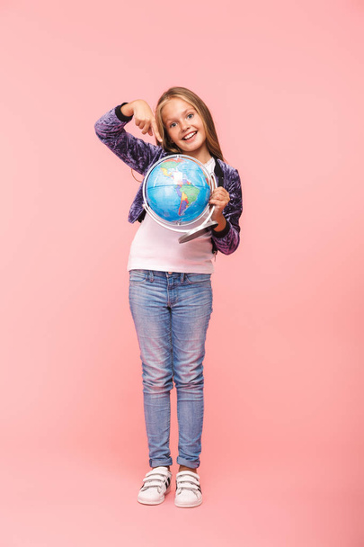 Full length of a smiling little girl standing isolated over pink background, holding earth globe - Valokuva, kuva