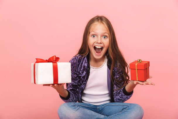 Excited pretty little girl sitting isolated over pink background, choosing between small and big gift boxes - Фото, изображение