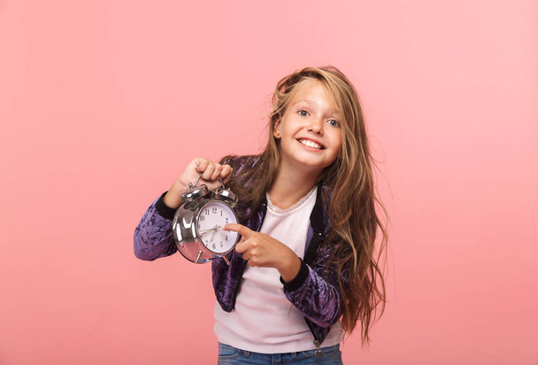 Pretty little girl sitting isolated over pink background, holding alarm clock - Foto, immagini