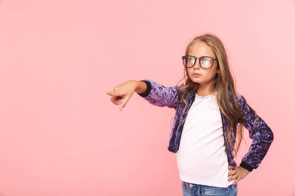 Portrait of a serious little girl wearing eyeglasses standing isolated over pink background, pointing at copy space - Fotó, kép