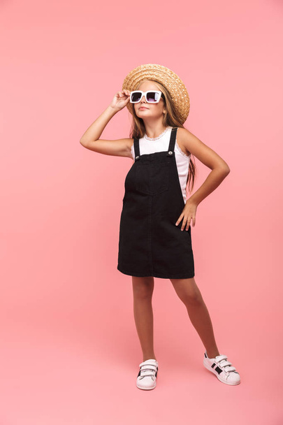 Full length portrait of a smiling little girl wearing summer sunglasses and hat isolated over pink background - Foto, immagini